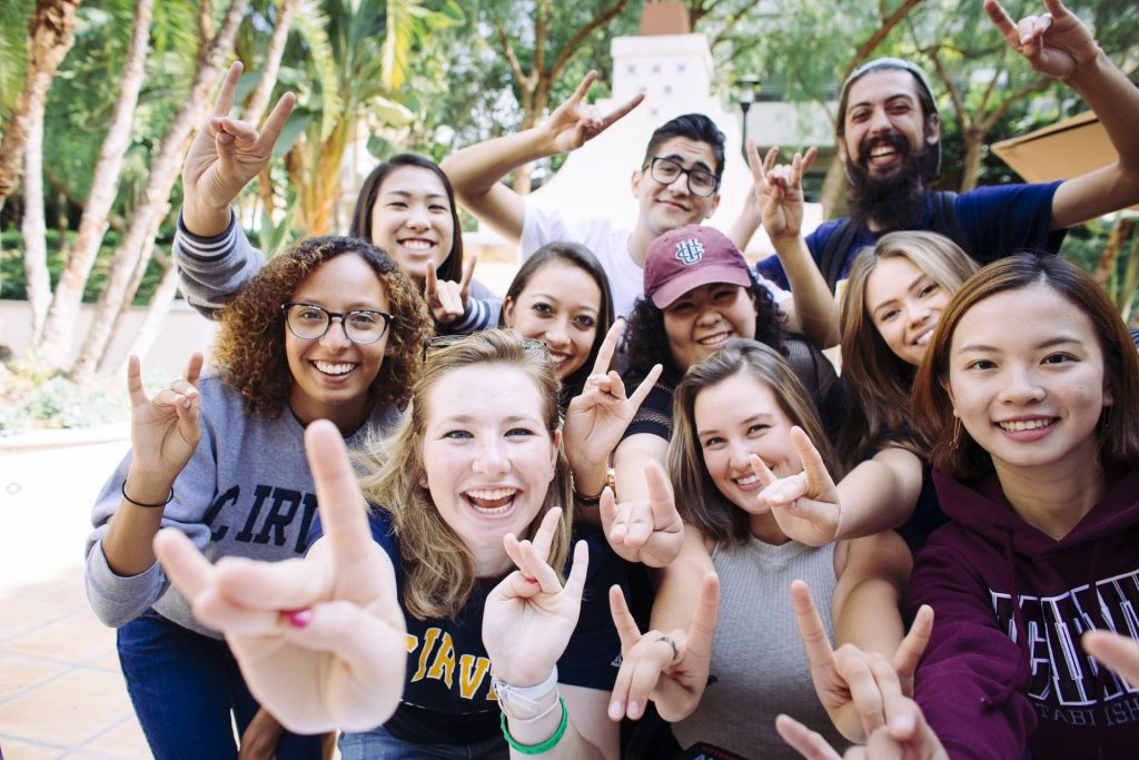 An image of 10 UCI students, smiling while wearing clothes with the UCI logo and making the anteater hand sign "Zot Zot" toward the camera. There are trees in the background.